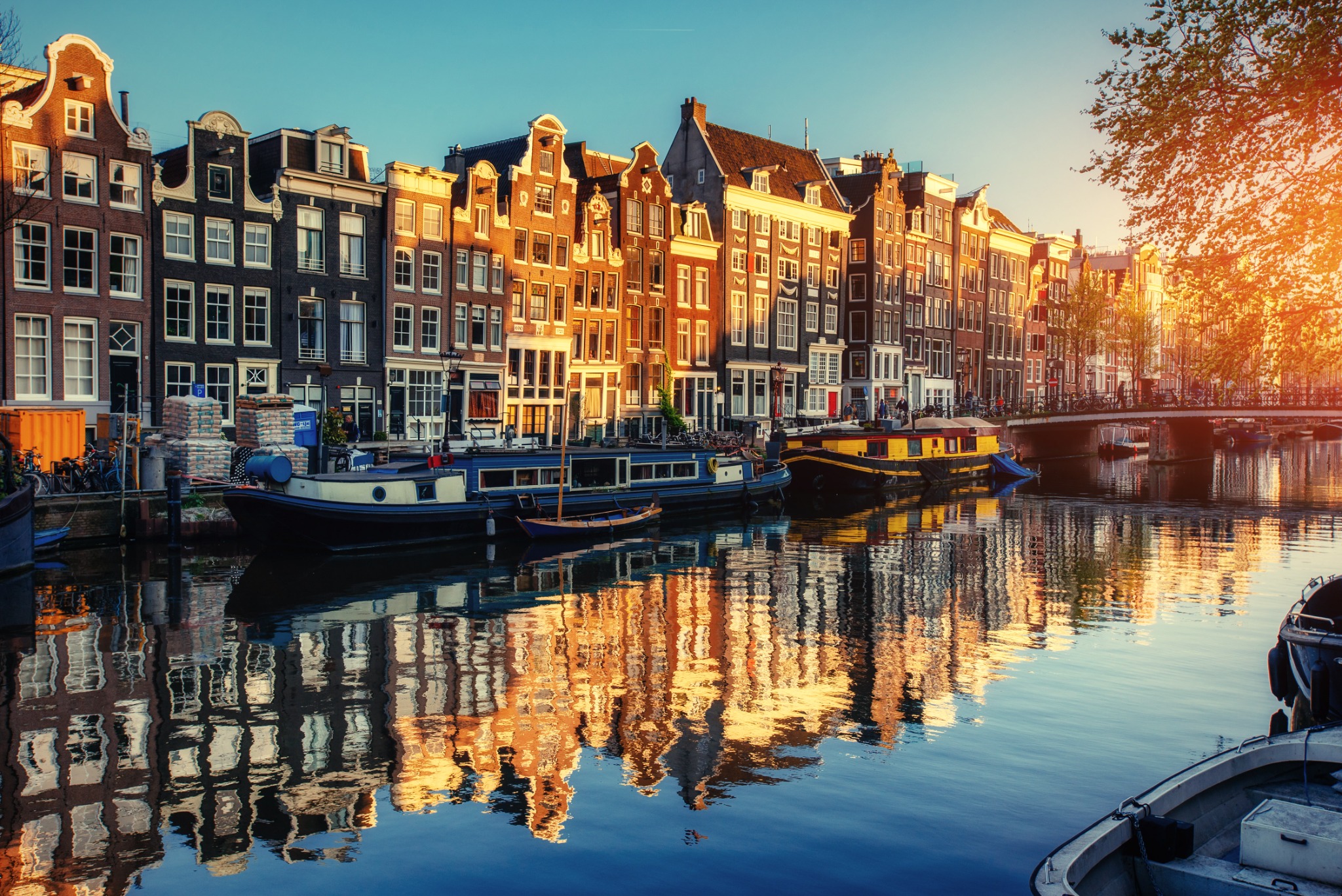 Golden sunset light illuminates Amsterdam canal houses, bridges, and moored boats.