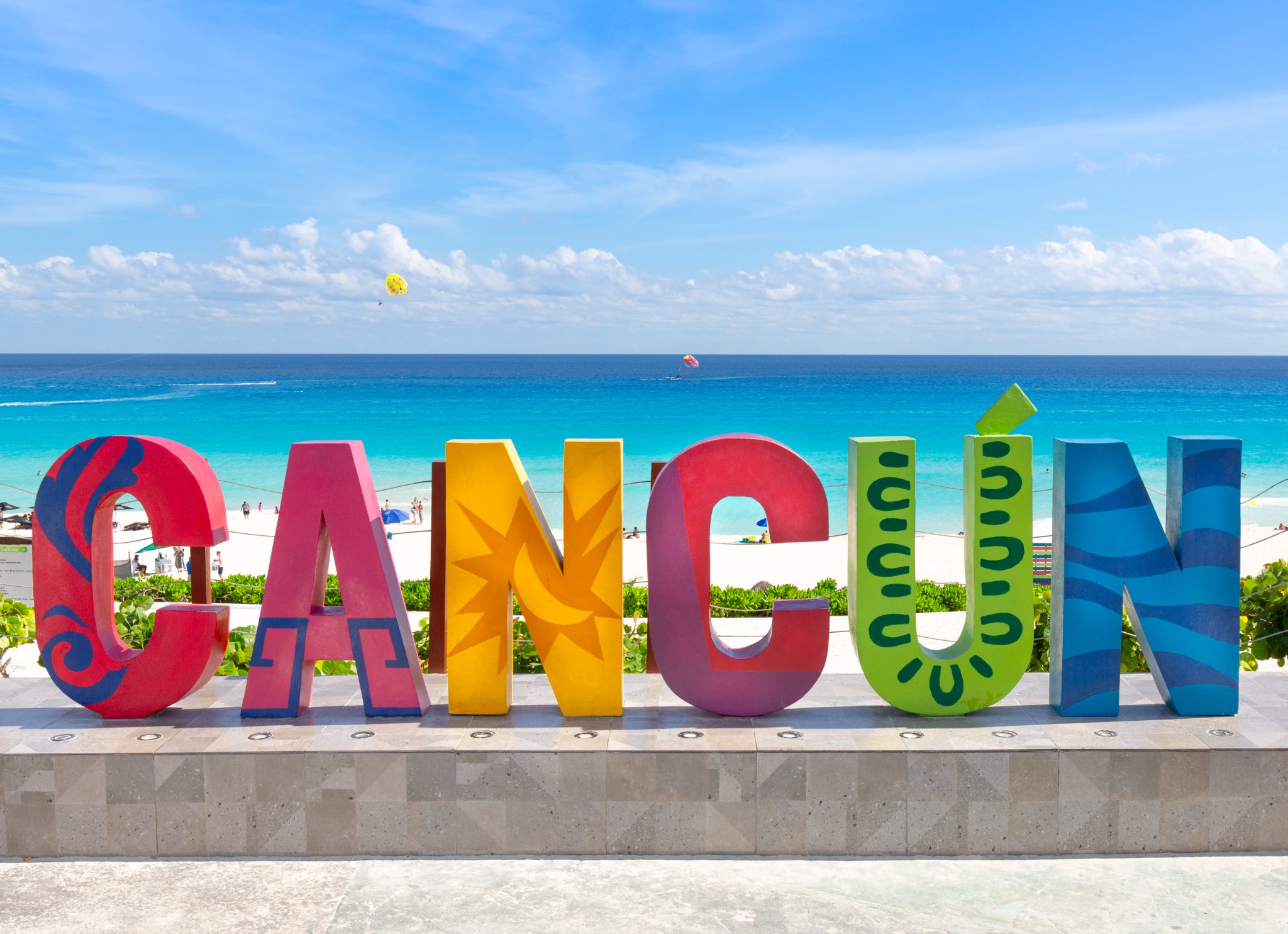 Bright travel photograph of the famous multicolored Cancún sign at Playa Delfines, with the Caribbean Sea and beach visible in the background.