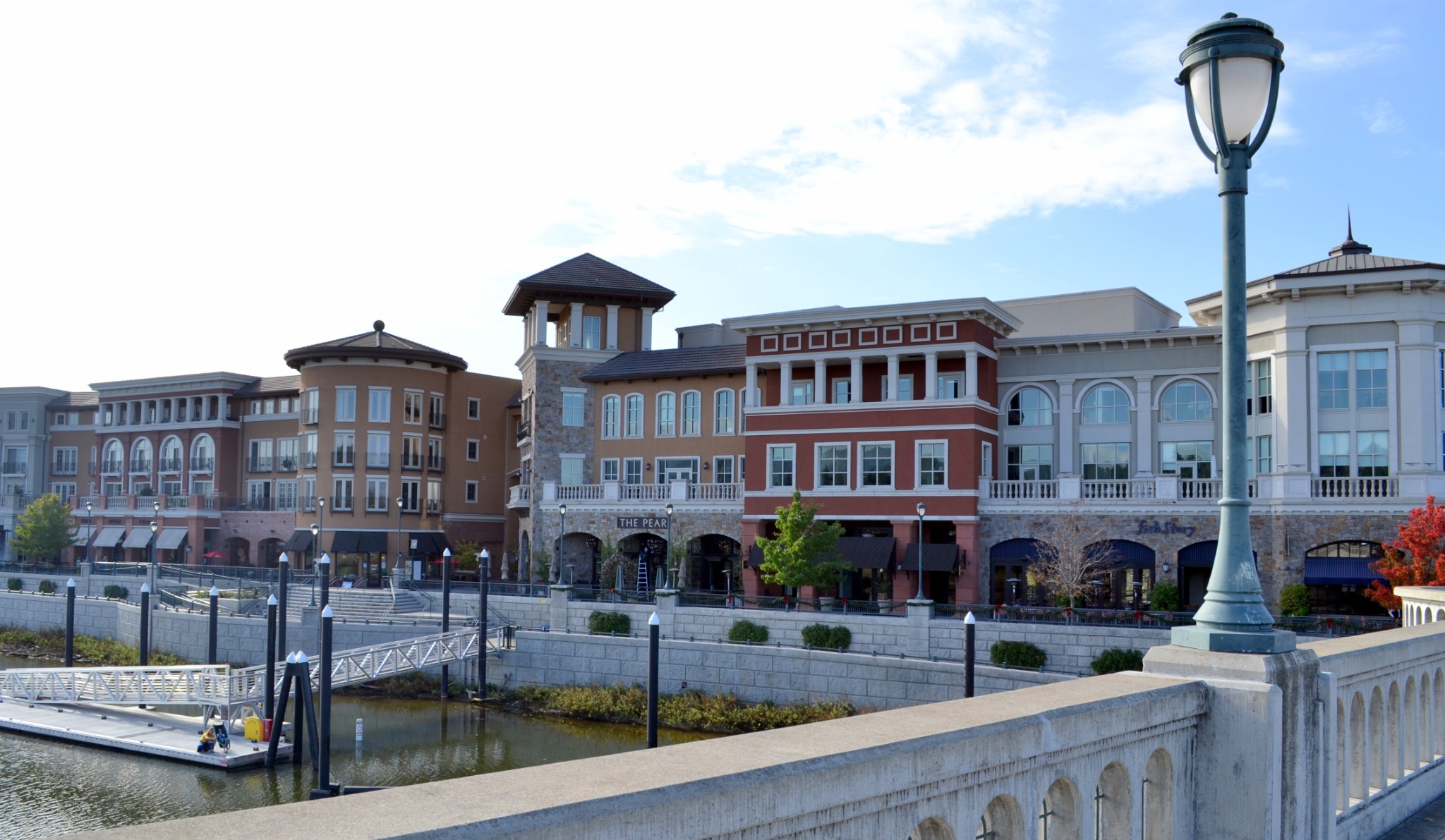 A daytime view of the Napa Riverfront in downtown Napa, featuring colorful buildings, restaurants, a marina, and pedestrian-friendly waterfront scenery. Alternative text: Downtown Napa buildings and restaurants beside the Napa River in California. Title: Downtown Napa Riverfront Caption: The downtown Napa riverfront with shops, restaurants, and waterfront architecture. Description: A daytime view of the Napa Riverfront in downtown Napa, featuring colorful buildings, restaurants, a marina, and pedestrian-friendly waterfront scenery.