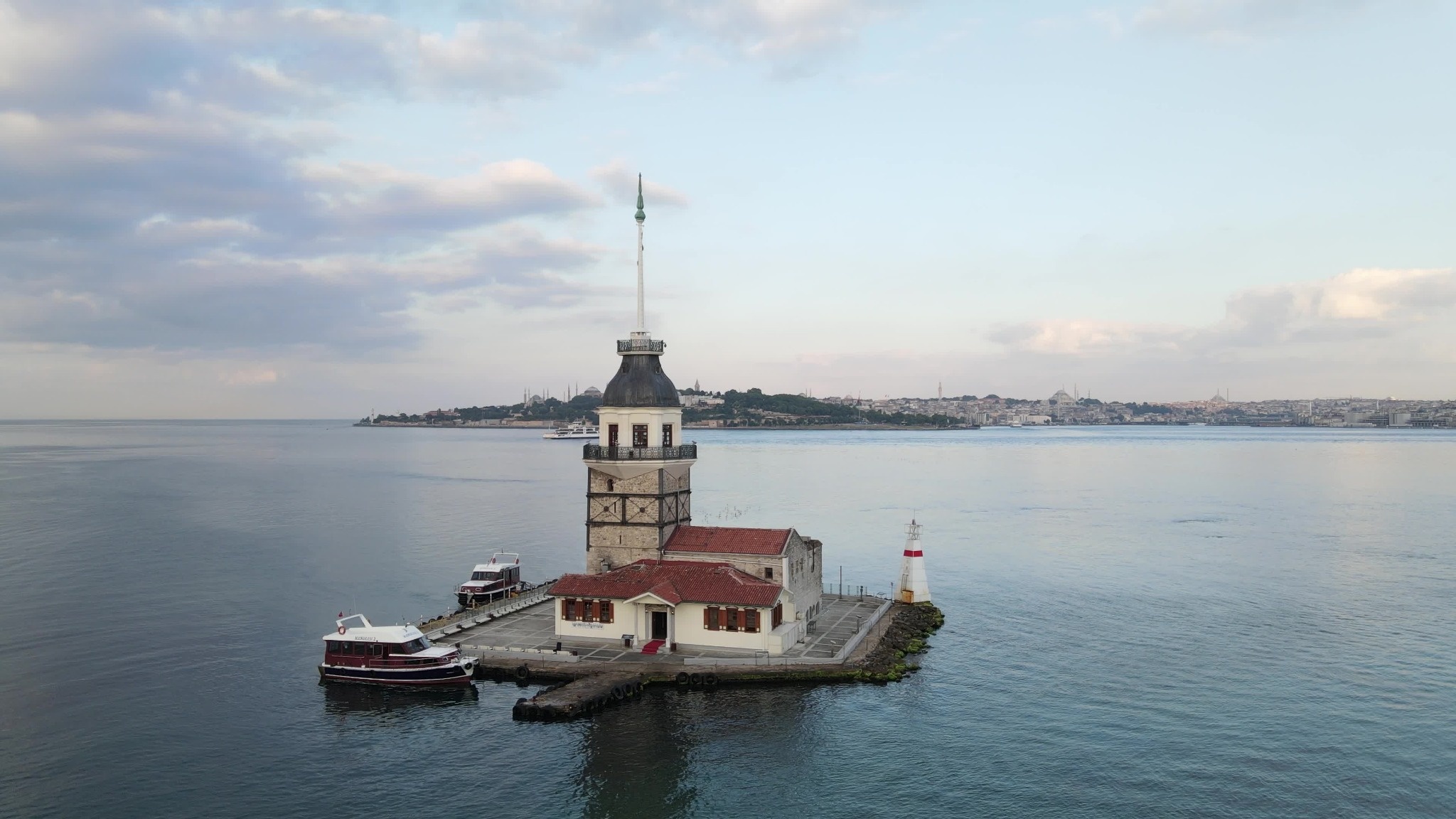 Aerial view of Maiden's Tower on a small islet in the Bosphorus with Istanbul's historic skyline in the distance. A wide aerial photograph of Maiden's Tower, also known as K?z Kulesi, surrounded by the Bosphorus in Istanbul, Turkey. Passenger boats sit beside the landmark while the historic city skyline stretches across the background.
