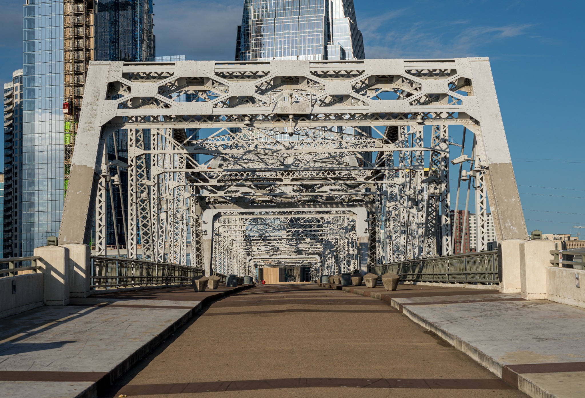 A symmetrical view through the steel framework of the John Seigenthaler Pedestrian Bridge, with modern downtown Nashville buildings rising behind it. Alternative text: John Seigenthaler Pedestrian Bridge in downtown Nashville framed by modern high-rise buildings. Title: John Seigenthaler Pedestrian Bridge in Nashville Caption: The steel structure of Nashville's John Seigenthaler Pedestrian Bridge in daylight. Description: A symmetrical view through the steel framework of the John Seigenthaler Pedestrian Bridge, with modern downtown Nashville buildings rising behind it. Keywords: Nashville, John Seigenthaler Pedestrian Bridge, downtown Nashville, Cumberland River, Tennessee
