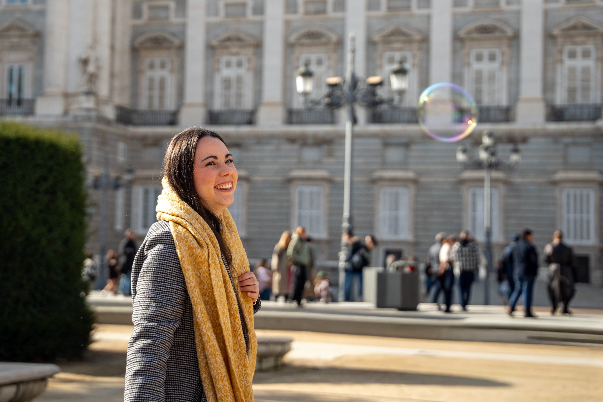 Smiling traveler watching a soap bubble outside the Royal Palace of Madrid on a sunny day.