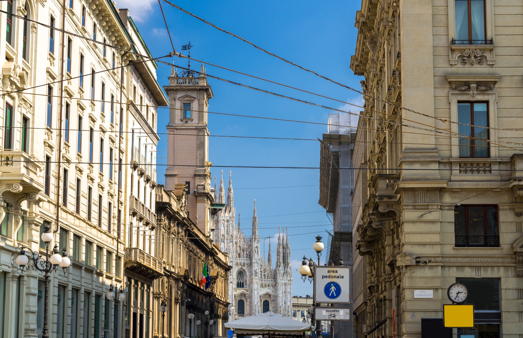 Daytime street scene in central Milan with elegant architecture, overhead tram lines, and the spires of the Duomo di Milano visible in the distance.