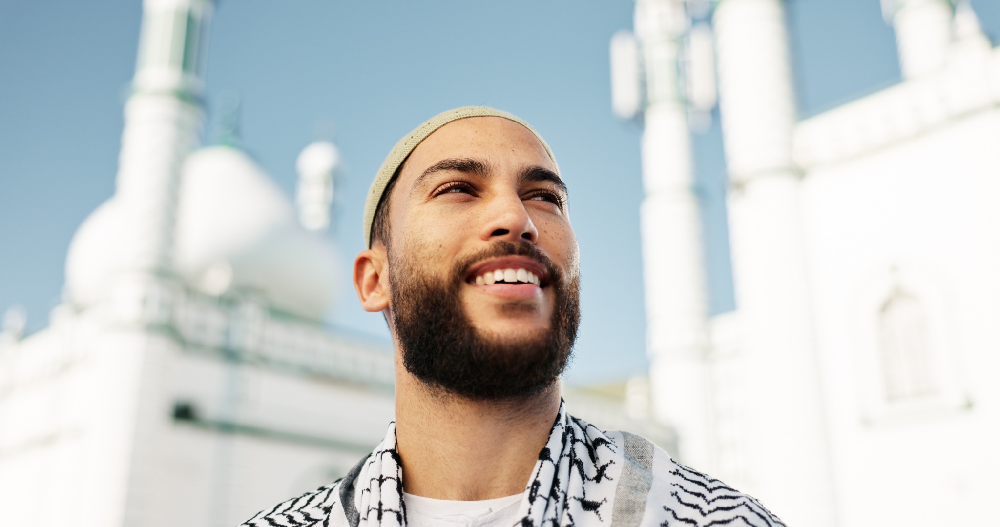 Alternative text: Smiling Muslim man wearing a prayer cap outside a mosque in Mecca. Title: Muslim Visitor Outside a Mosque in Mecca Caption: A smiling Muslim visitor stands outside a mosque in Mecca under a clear blue sky. Description: Portrait of a smiling Muslim man wearing a prayer cap and patterned scarf outside a mosque in Mecca, Saudi Arabia. Mosque domes and minarets appear softly blurred in the background. Portrait of a smiling Muslim man wearing a prayer cap and patterned scarf outside a mosque in Mecca, Saudi Arabia. Mosque domes and minarets appear softly blurred in the background.