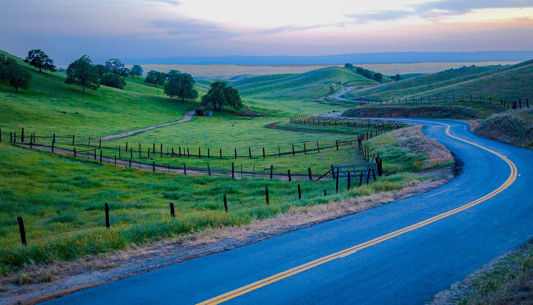 A peaceful two-lane country road curves through green hills, oak trees, and ranchland in the broader Northern California wine-country landscape near Napa Valley. Alternative text: Winding country road crossing rolling green hills in California wine country. Title: Scenic Country Road Near Napa Valley Caption: A winding rural road through the rolling hills of Northern California wine country. Description: A peaceful two-lane country road curves through green hills, oak trees, and ranchland in the broader Northern California wine-country landscape near Napa Valley.