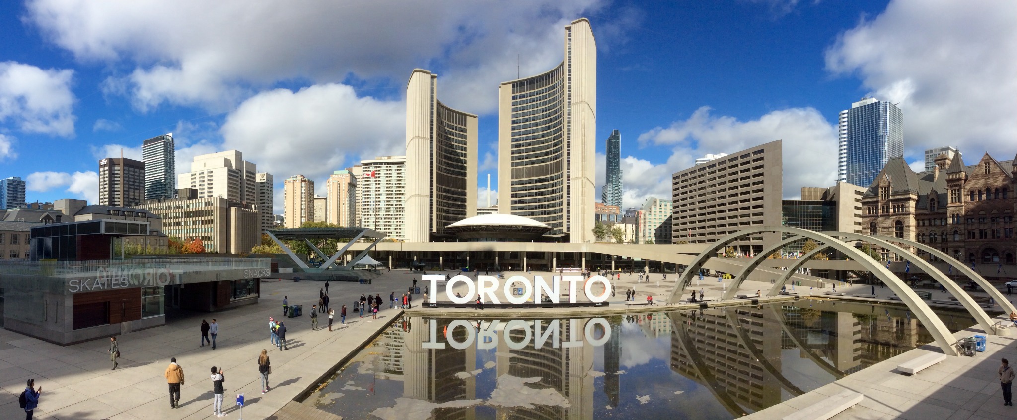 A panoramic view of Nathan Phillips Square in downtown Toronto, including the Toronto sign, reflecting pool, curved City Hall towers and surrounding skyline. The square is a central civic gathering place.