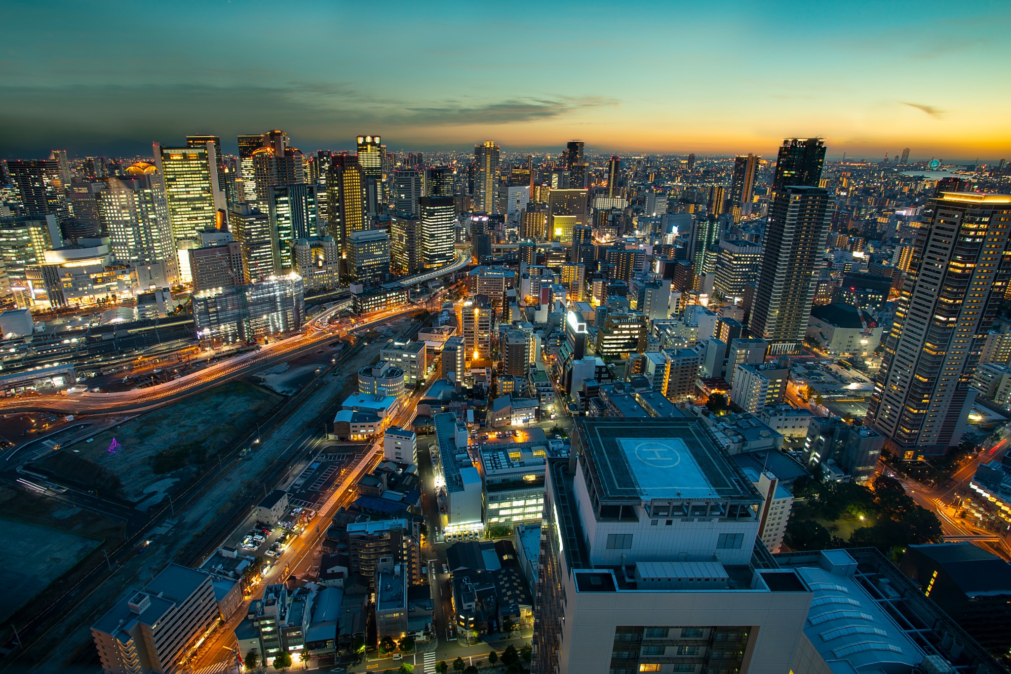 A sweeping high-angle panorama of central Osaka at sunset, featuring illuminated skyscrapers, rail lines, roads and dense city blocks extending toward the horizon.