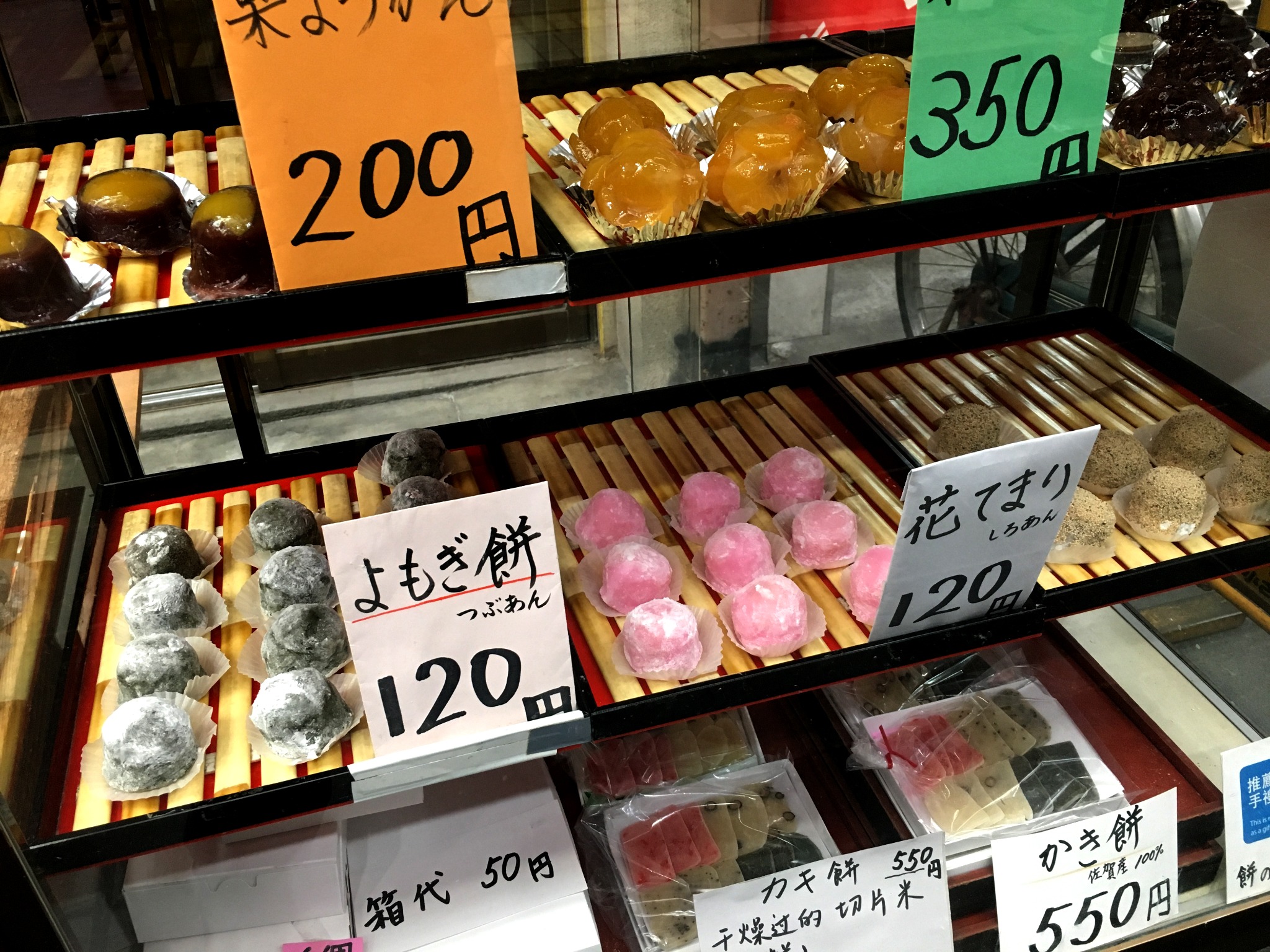 A shop display filled with pink, green, white and brown Japanese mochi sweets, accompanied by handwritten labels and prices in yen. The image highlights Osaka's everyday food culture.