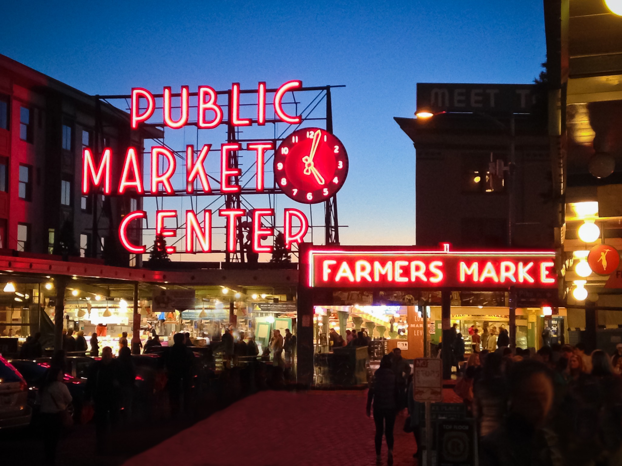 The iconic red neon Public Market Center sign and clock illuminate the busy entrance to Pike Place Market in downtown Seattle during the evening.
