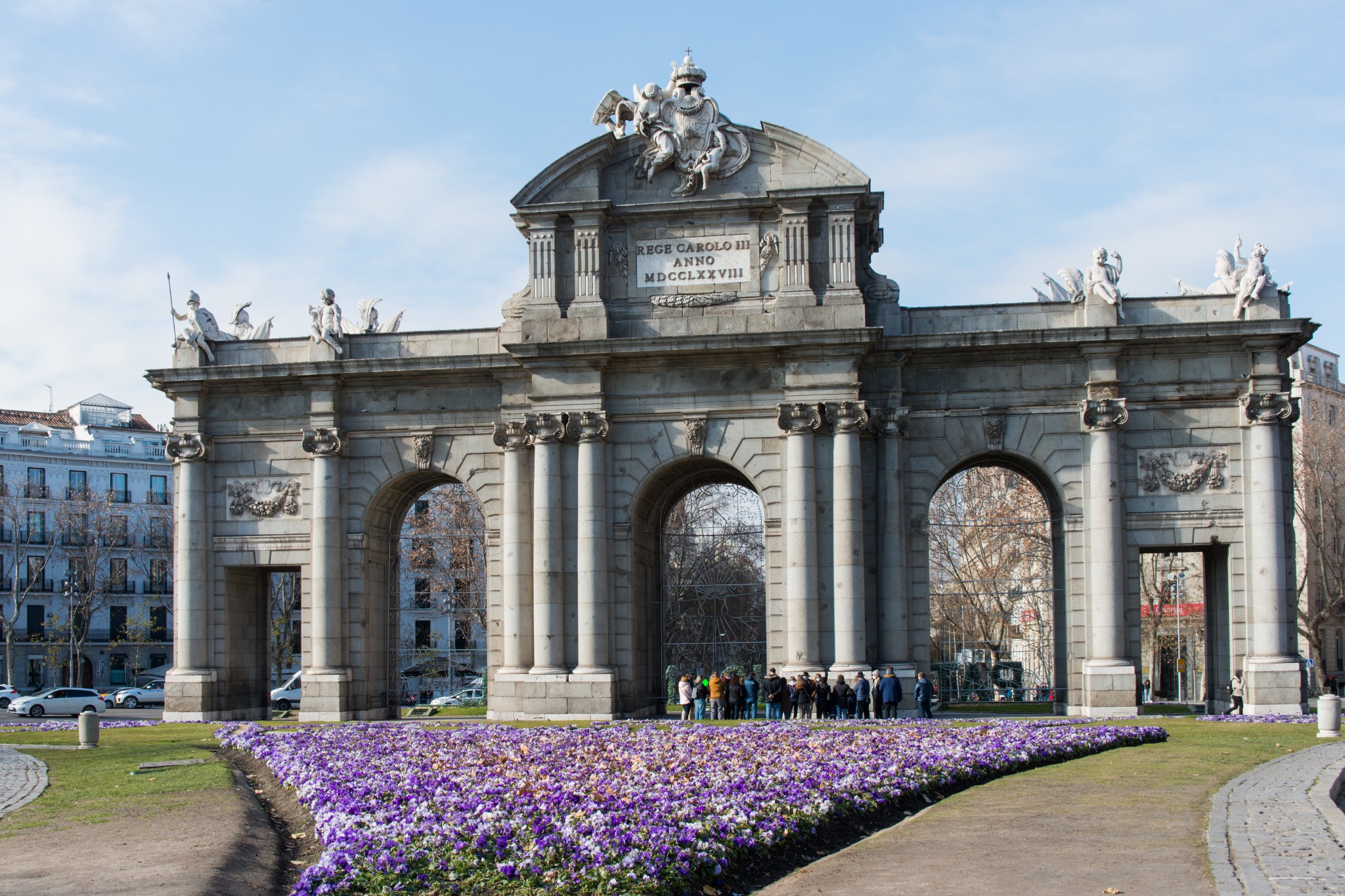 Puerta de Alcala monument in Madrid framed by purple flowers on a sunny day.