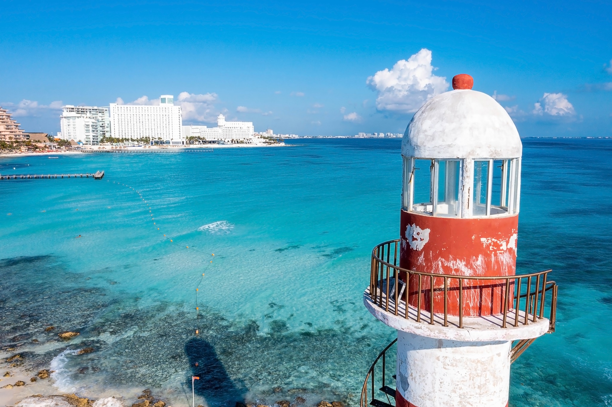 Scenic aerial-style view of the Punta Cancún lighthouse above clear turquoise water, with hotels and the Cancún coastline in the distance.