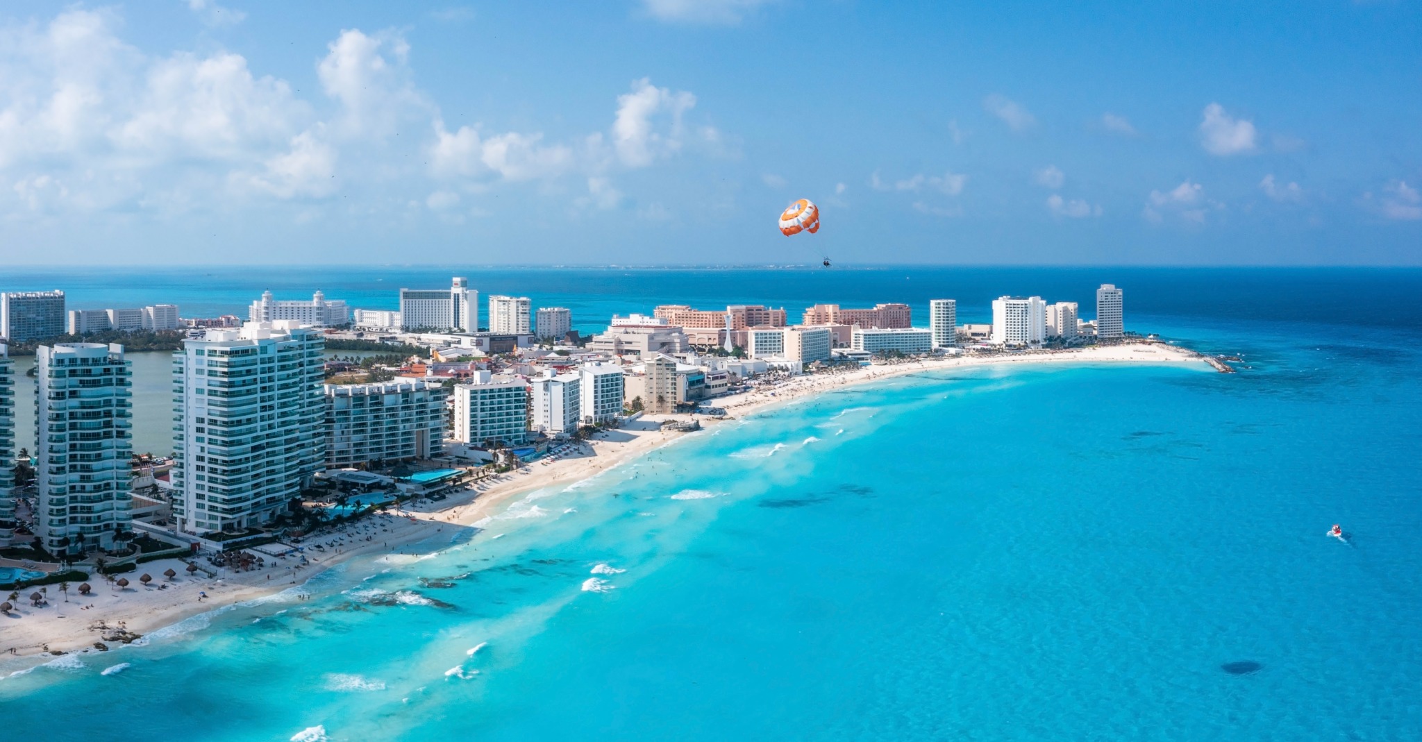 Panoramic aerial travel image of the Punta Norte coastline near Cancún, with beachfront resorts, white sand, vivid turquoise water and parasailing offshore.