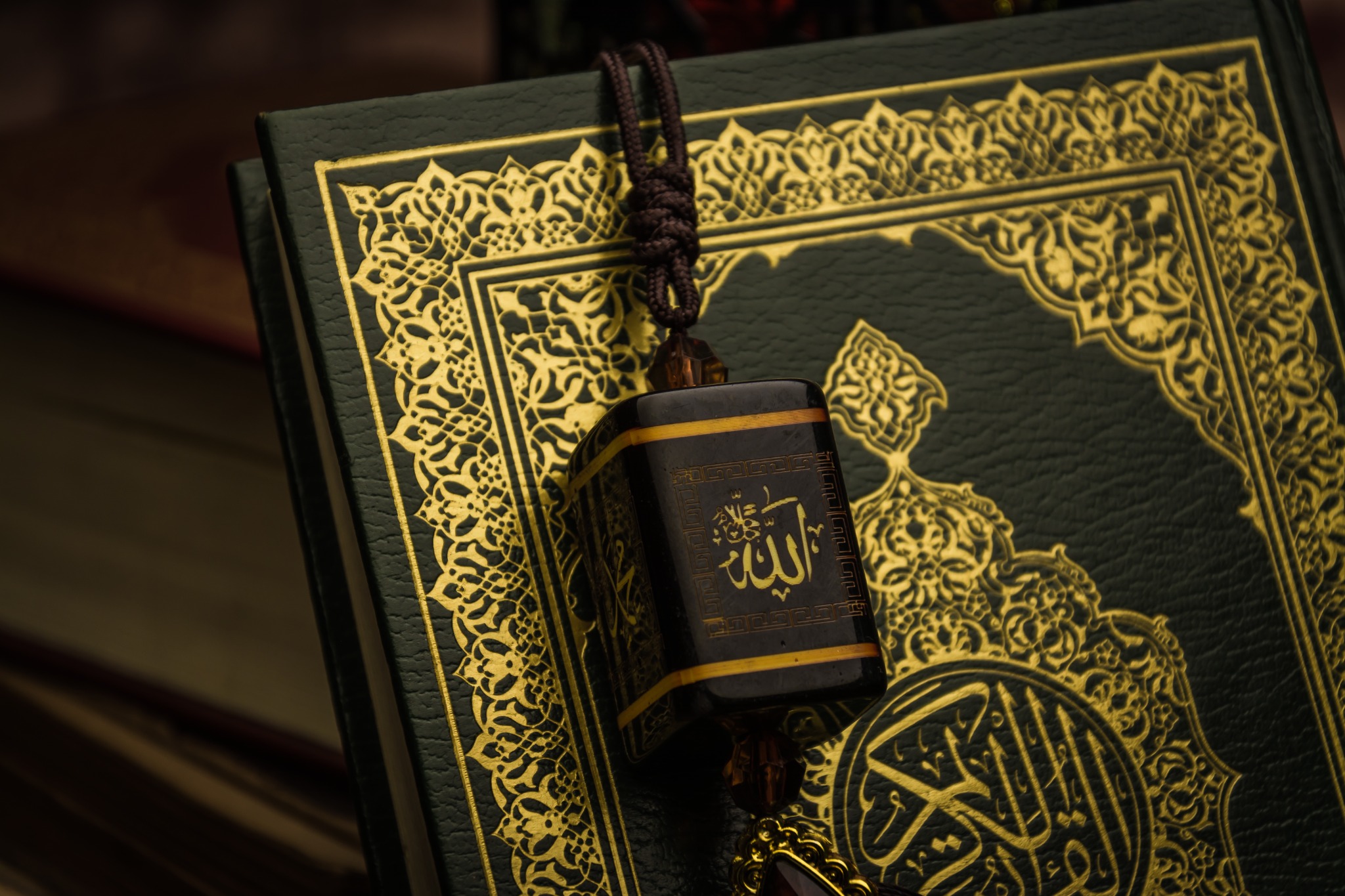 Alternative text: Decorative Quran cover with a pendant bearing the name Allah in Arabic calligraphy. Title: Quran with Allah Calligraphy Pendant Caption: An ornate Quran and decorative pendant featuring the name Allah in Arabic calligraphy. Description: Close-up still life of an ornate green Quran with gold decoration and a hanging pendant bearing the name Allah in Arabic calligraphy. Warm, subdued lighting draws attention to the intricate religious artwork. Close-up still life of an ornate green Quran with gold decoration and a hanging pendant bearing the name Allah in Arabic calligraphy. Warm, subdued lighting draws attention to the intricate religious artwork.