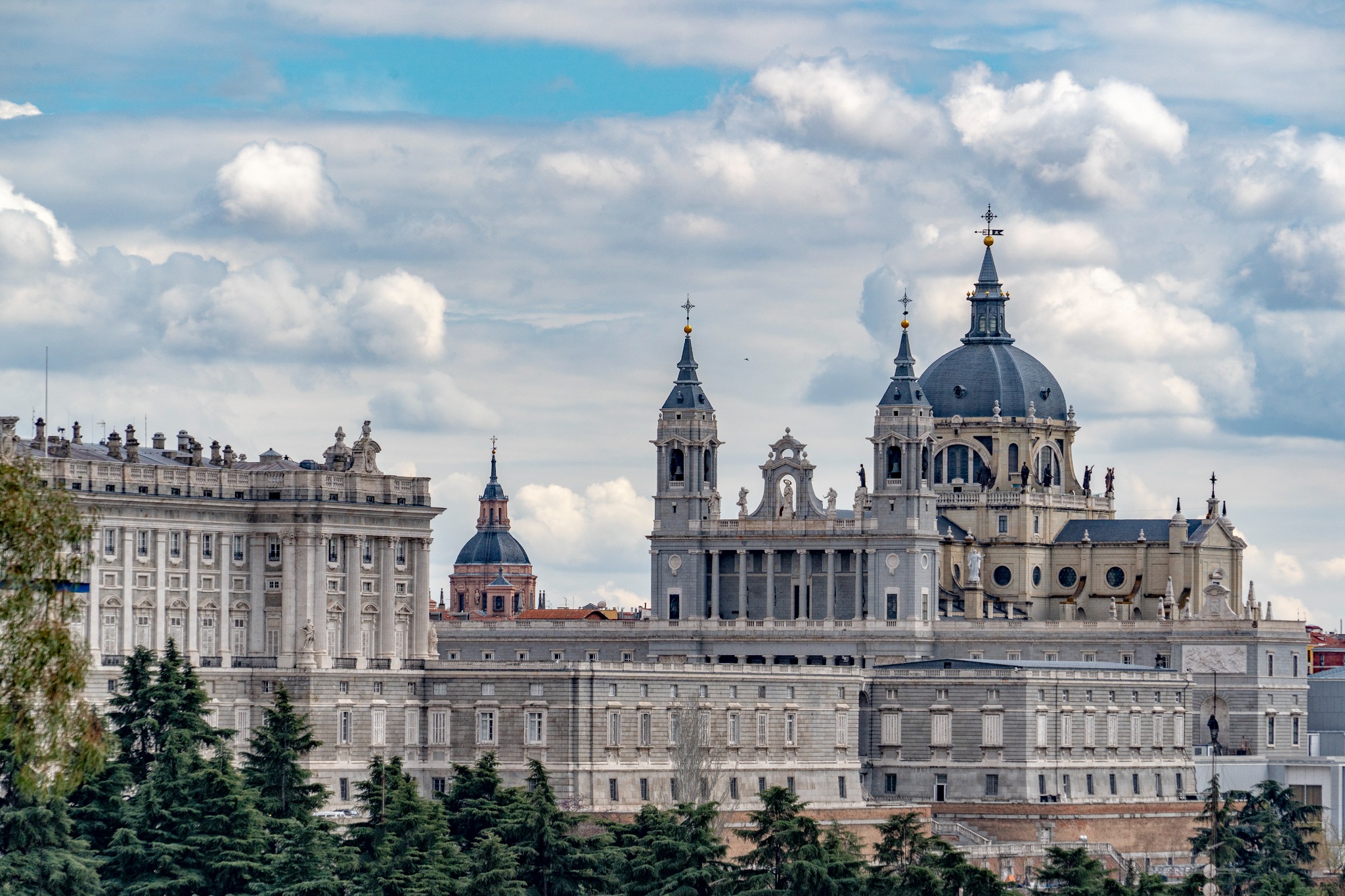 Royal Palace of Madrid and Almudena Cathedral rising above trees under a dramatic cloudy sky.