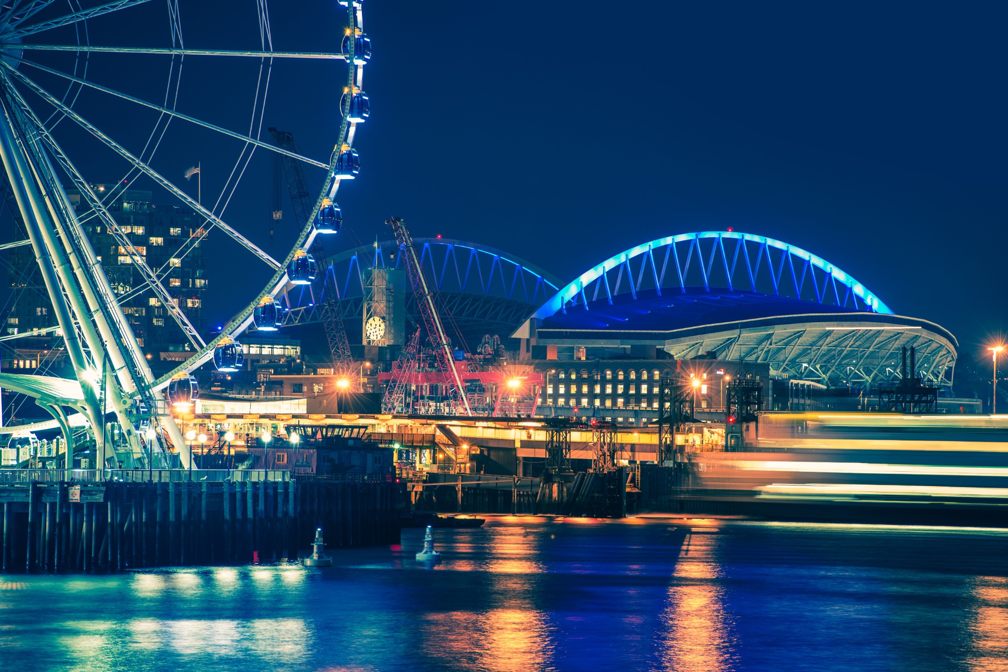 A vivid nighttime view of the Seattle waterfront featuring the illuminated Seattle Great Wheel, Lumen Field, waterfront buildings, construction cranes, and a passing ferry reflected in Elliott Bay.