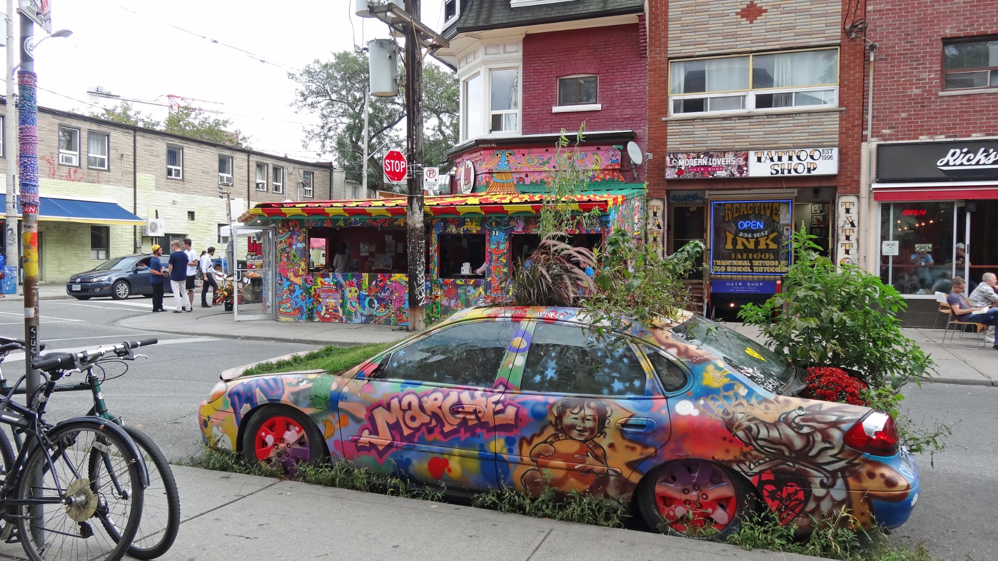 A colorful street scene in Kensington Market, Toronto, featuring a graffiti-covered car, vivid murals and independent storefronts. Kensington Market is known for its eclectic art, food and multicultural character.
