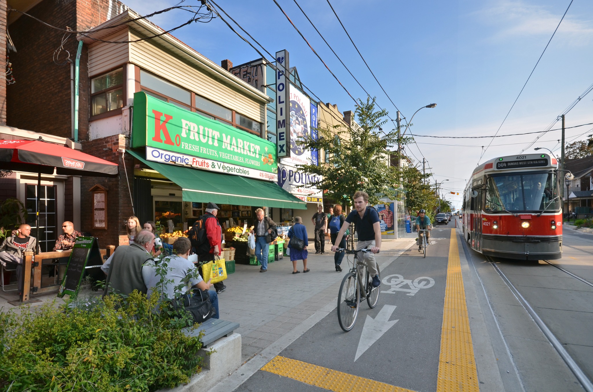 Street life on Roncesvalles Avenue in Toronto, with a TTC streetcar, neighborhood shops, cyclists and pedestrians. The image highlights the walkable character of Roncesvalles Village.