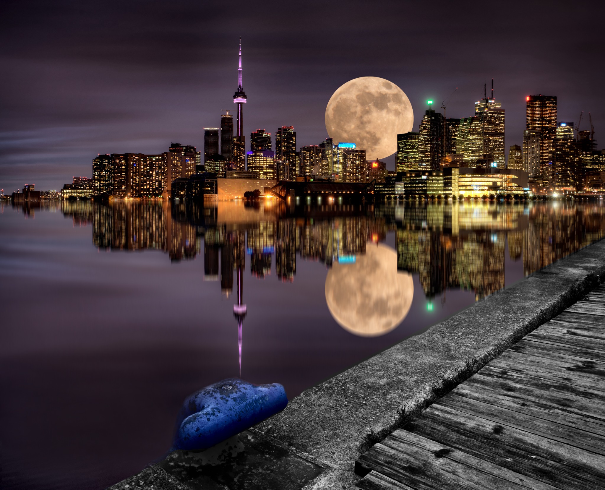 A dramatic nighttime view of the Toronto skyline with a large full moon, the CN Tower and city lights reflected on calm water. The scene presents Toronto's waterfront at its most atmospheric.