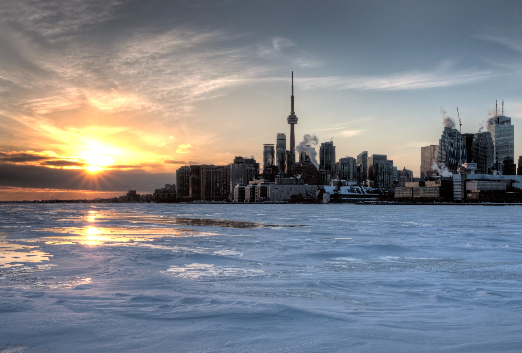 A winter view of the Toronto skyline from Polson Pier, with the CN Tower silhouetted against a golden sunset and ice covering the harbor. Polson Pier offers a wide panorama of downtown Toronto.