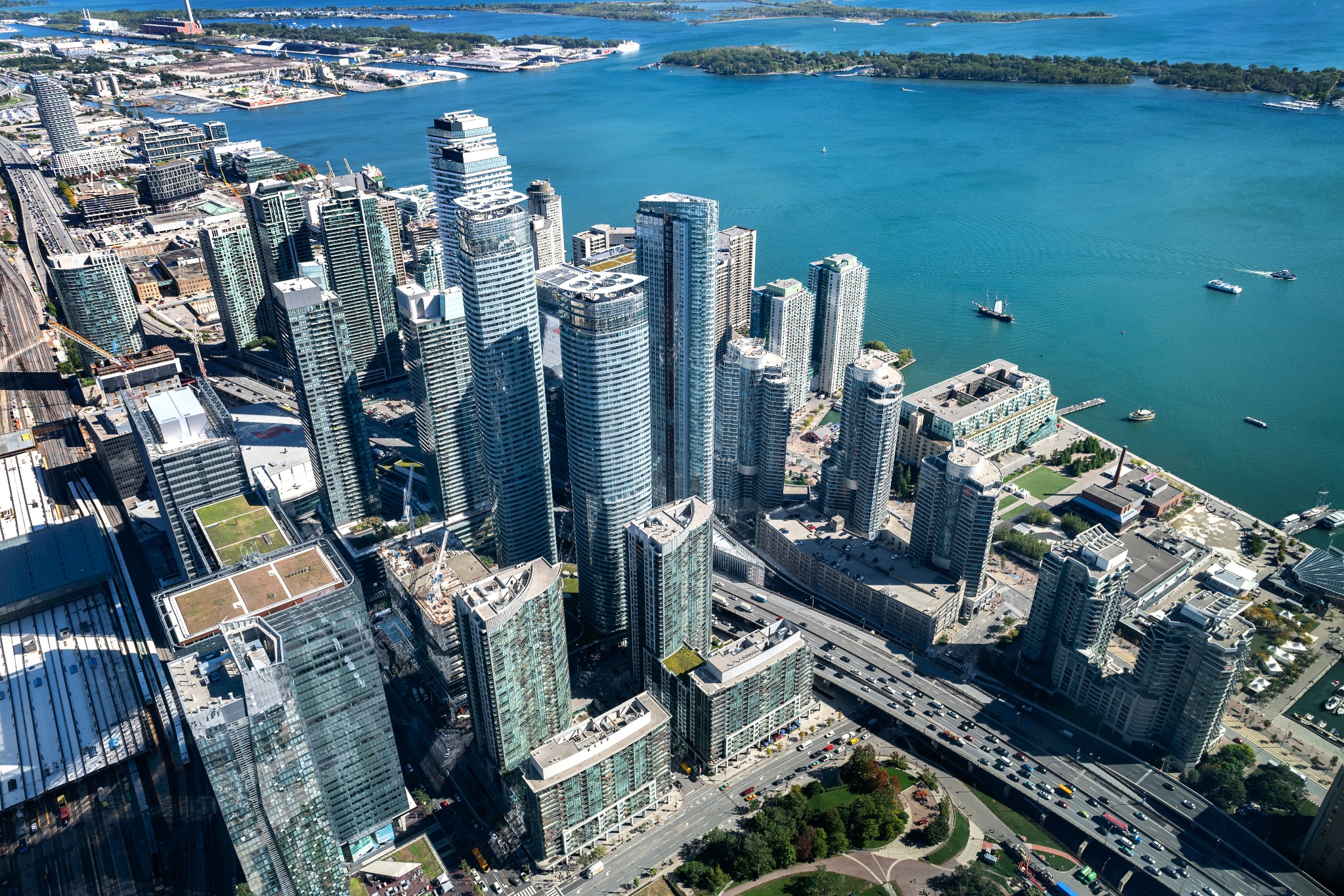 A high-angle view of Toronto's CityPlace neighborhood beside Lake Ontario, with condominium towers, the Gardiner Expressway, rail corridor and Toronto Islands in the distance. The image shows the density of the downtown waterfront.