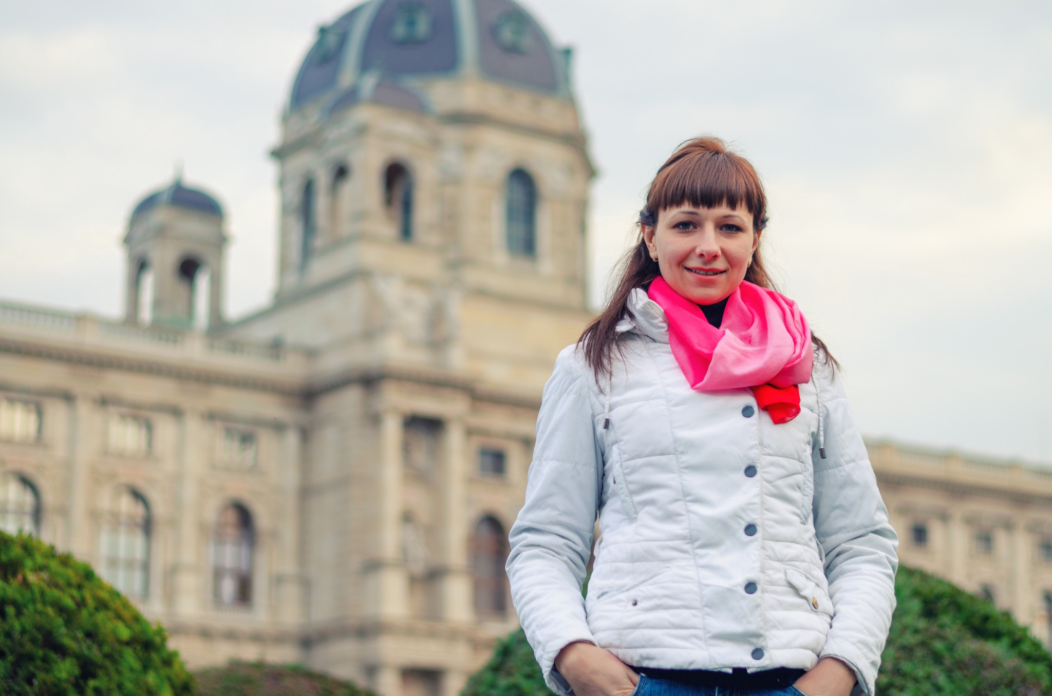 Travel image of a woman wearing a white jacket and bright pink scarf in front of a domed historic museum building in Vienna, Austria.